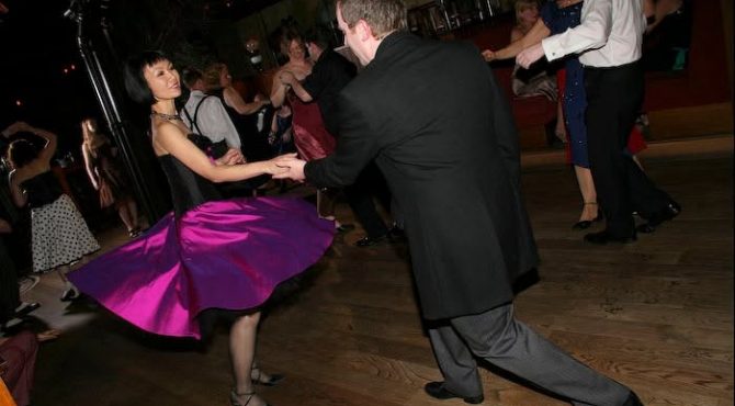 Joo-Lee in a swirling purple dress, dancing Lindy Hop with a partner on a wooden dance floor.